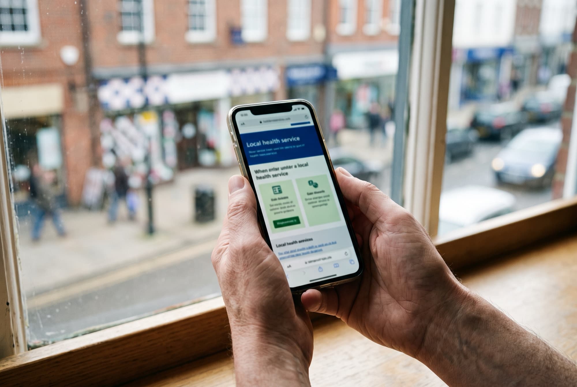 Someone checking a health service website on their phone, by a window looking out on a high street